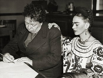 Frida Kahlo places her hand on Diego Rivera's shoulder as he signs paperwork in a black and white photo