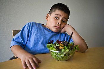 A preteen overweight boy sits at a table disappointed by the salad inf front of him