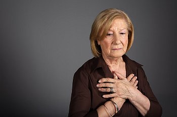 An older woman stands with hands over chest on gray background