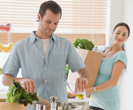A husband cooking dinner while his wife holds grocery bag and watches him